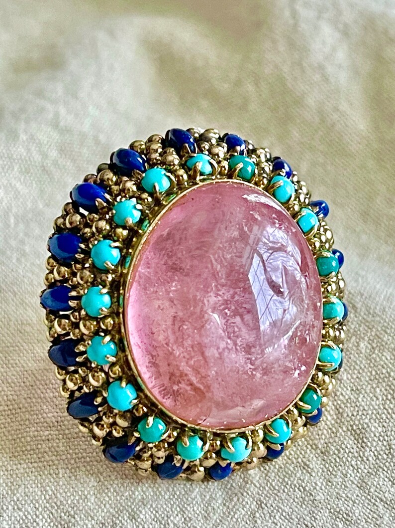 May include: A pink gemstone ring with a gold setting and a halo of blue and turquoise stones.
