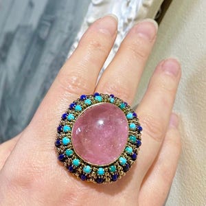 May include: A large pink gemstone ring with a gold setting. The ring is surrounded by a halo of blue and turquoise stones.