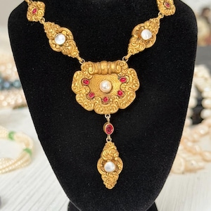 May include: Ornate gold-toned necklace with a central pendant and a series of connecting links. The pendant and links feature red and white accents. The necklace is displayed on a black velvet bust.