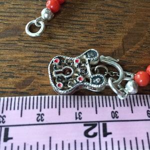 Puede incluir: Un collar de cuentas de coral rojo con un cierre de plata en forma de candado. El candado tiene detalles de esmalte rojo y una cadena unida al cierre.