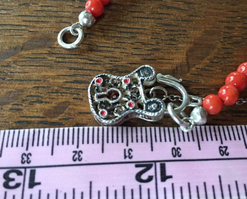 Puede incluir: Un collar de cuentas de coral rojo con un cierre de plata en forma de candado. El candado tiene detalles de esmalte rojo y una cadena unida al cierre.