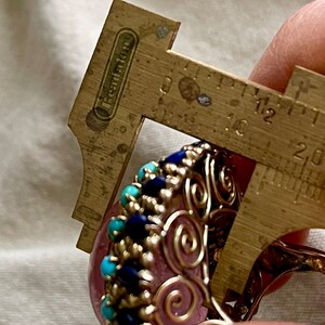 May include: A pink gemstone ring with a silver wire design and blue and turquoise stones. The ring is being measured with a caliper, showing a size of 1.8 cm.