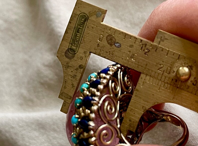 May include: A pink gemstone ring with a silver wire design and blue and turquoise stones. The ring is being measured with a caliper, showing a size of 1.8 cm.