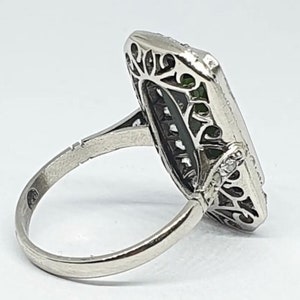 May include: A silver ring with an elongated rectangular face. The face features a central green stone, surrounded by filigree and small clear stones. The band is smooth and polished.