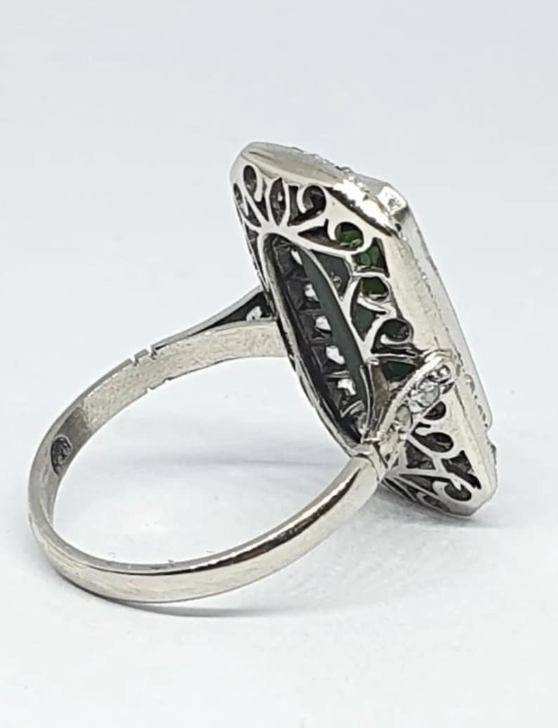 May include: A silver ring with an elongated rectangular face. The face features a central green stone, surrounded by filigree and small clear stones. The band is smooth and polished.