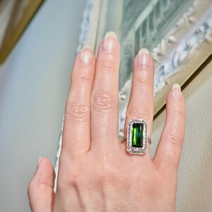 May include: A hand wearing a rectangular emerald-cut green gemstone ring with a halo of small white gemstones. The ring is set in a silver or white gold band. The ring is a piece of jewelry.