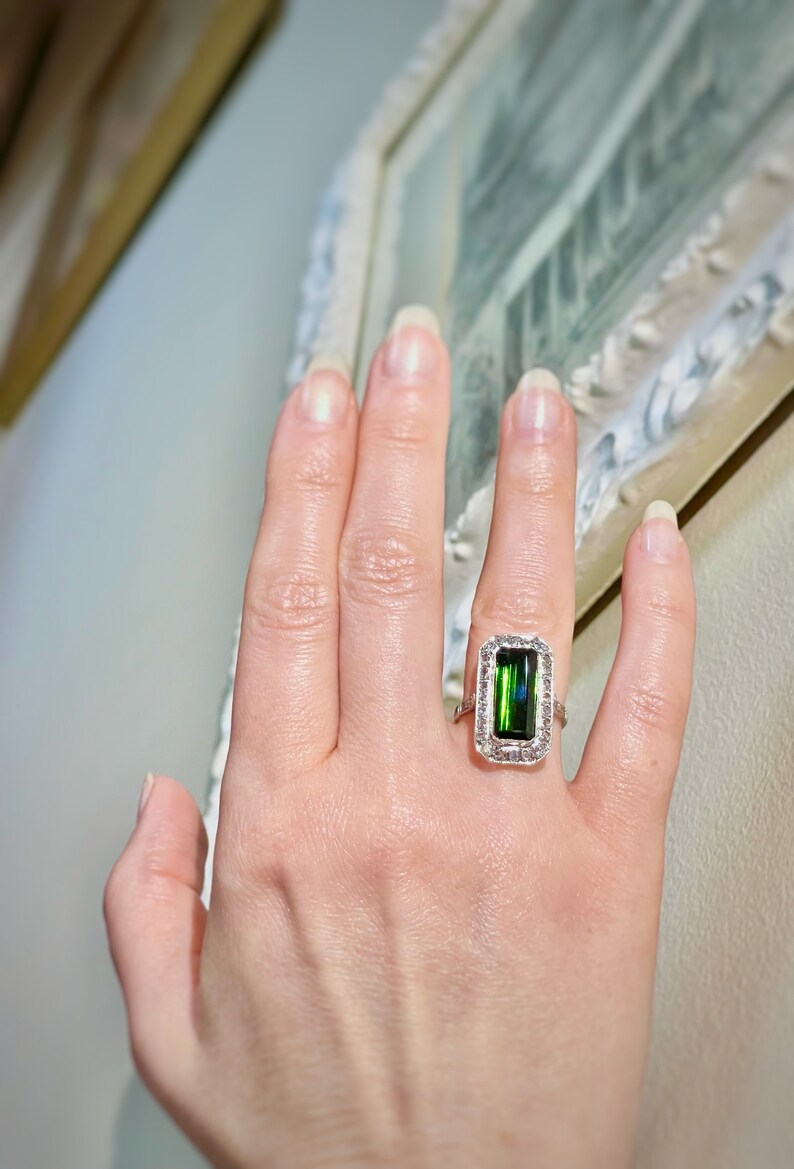 May include: A hand wearing a rectangular emerald-cut green gemstone ring with a halo of small white gemstones. The ring is set in a silver or white gold band. The ring is a piece of jewelry.