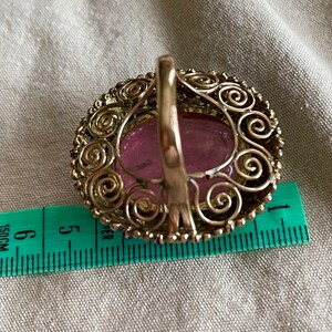 May include: A silver ring with a pink oval stone set in a filigree design. The ring is 1.5 inches in diameter.