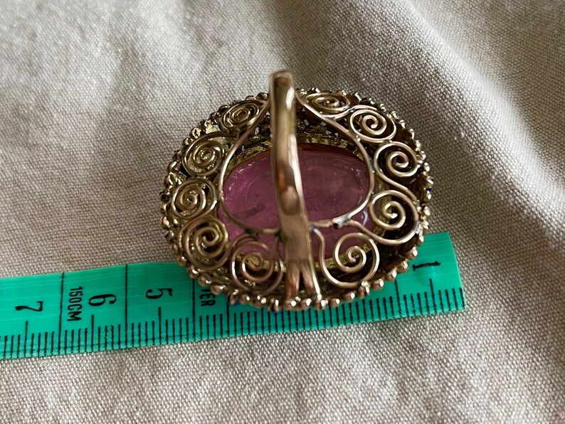 May include: A silver ring with a pink oval stone set in a filigree design. The ring is 1.5 inches in diameter.