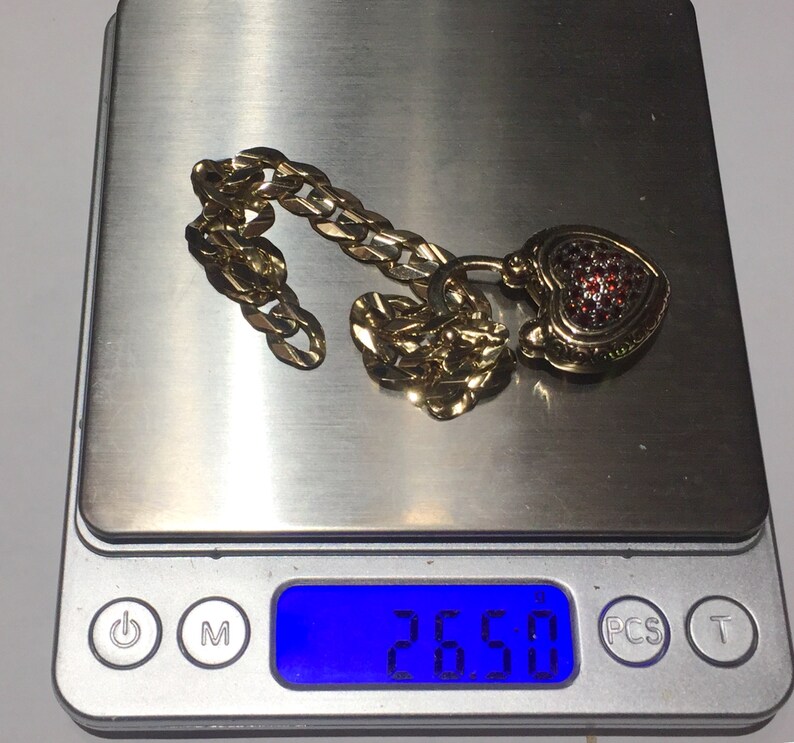 May include: A gold chain necklace with a heart-shaped pendant encrusted with red gemstones. The necklace is on a digital scale that reads 26.50.