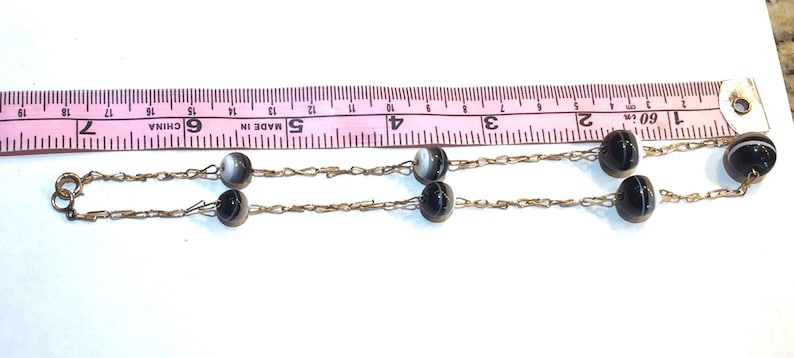 May include: A vintage necklace featuring alternating black and white striped beads strung on a delicate gold-tone chain. The necklace is shown with a pink measuring tape.