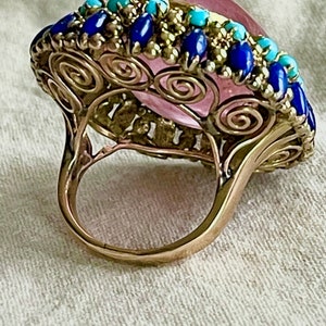 May include: A gold ring with a large pink gemstone surrounded by a border of blue and turquoise stones.