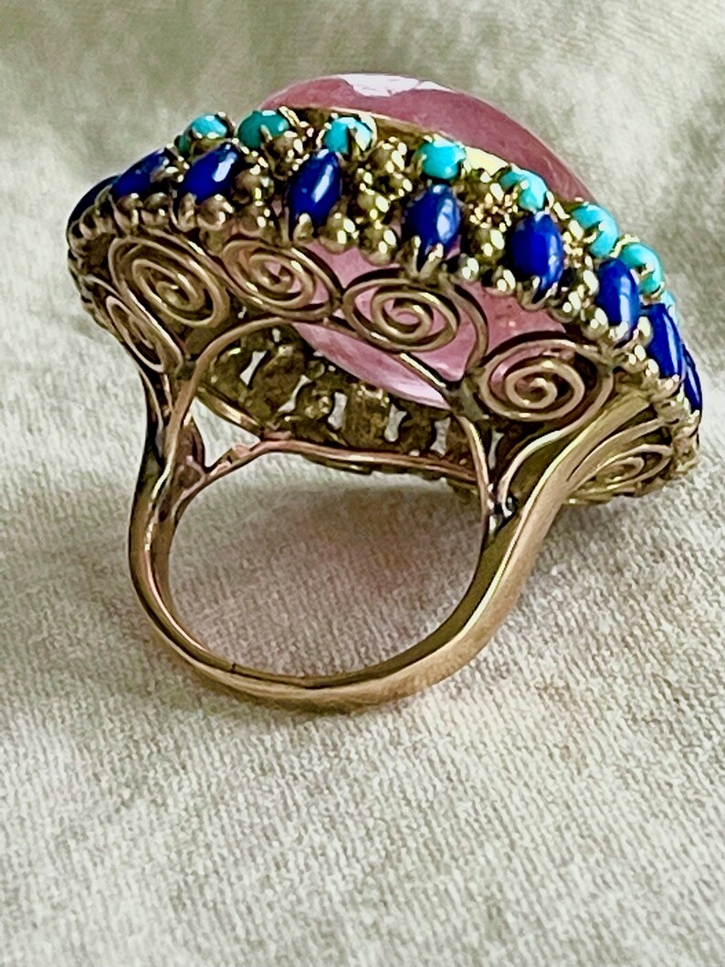 May include: A gold ring with a large pink gemstone surrounded by a border of blue and turquoise stones.