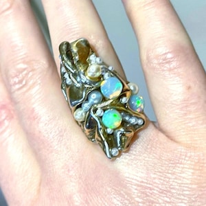Könnte beinhalten: Ein breiter, texturierter goldfarbener Ring mit einer organischen, geschmolzenen Metalloberfläche. Der Ring ist mit mehreren runden, irisierenden Opalen besetzt, die grüne und blaue Blitze zeigen, durchsetzt mit zahlreichen kleinen, weißen Perlen.