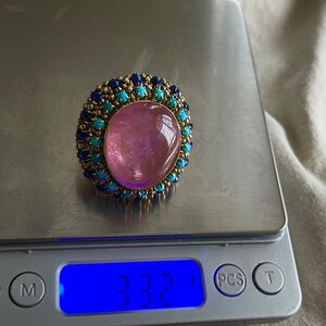 May include: A pink gemstone ring with a gold setting and a halo of blue and turquoise stones. The ring is on a digital scale that reads 3.32.