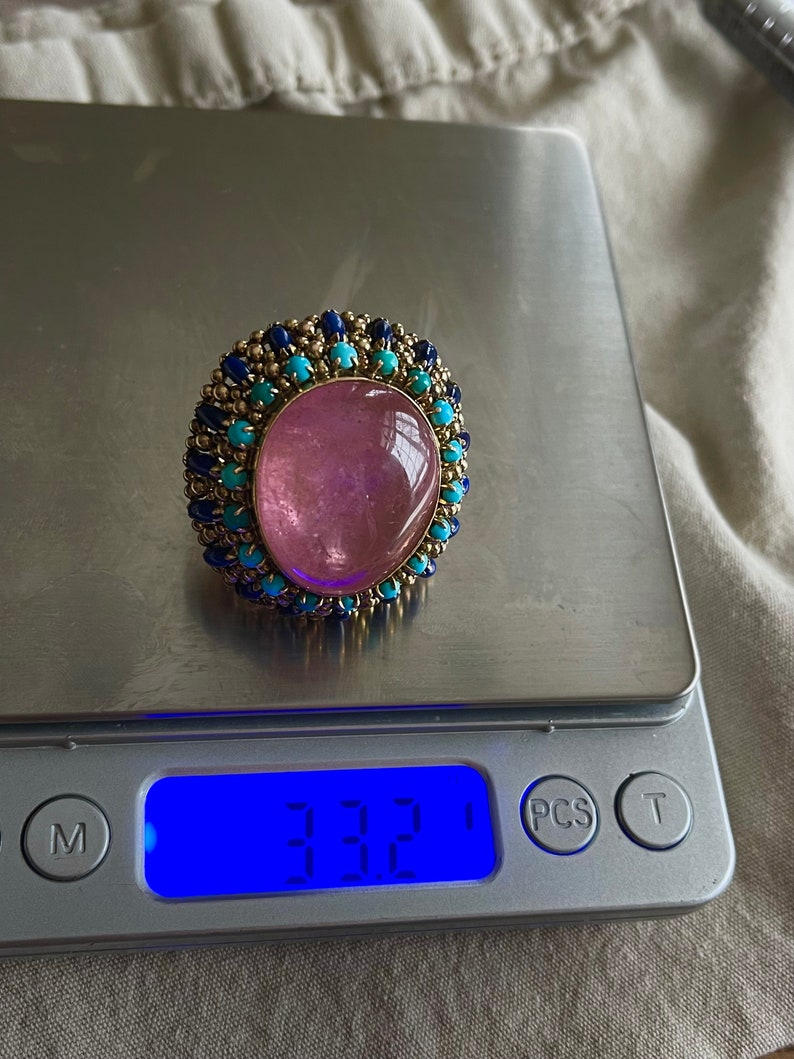 May include: A pink gemstone ring with a gold setting and a halo of blue and turquoise stones. The ring is on a digital scale that reads 3.32.