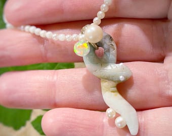 May include: A necklace featuring a strand of small white pearls, a larger cream-colored pearl, a small iridescent opal bead, and a carved light green stone snake pendant accented with two tiny white pearls near the tail of the snake figure.