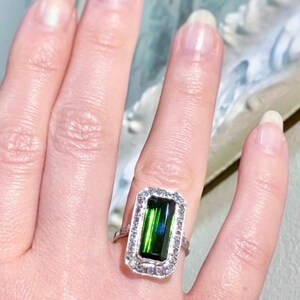 May include: A rectangular emerald-cut green gemstone ring with a halo of small, clear gemstones. The ring is set in a silver-colored band. The ring is on a finger.