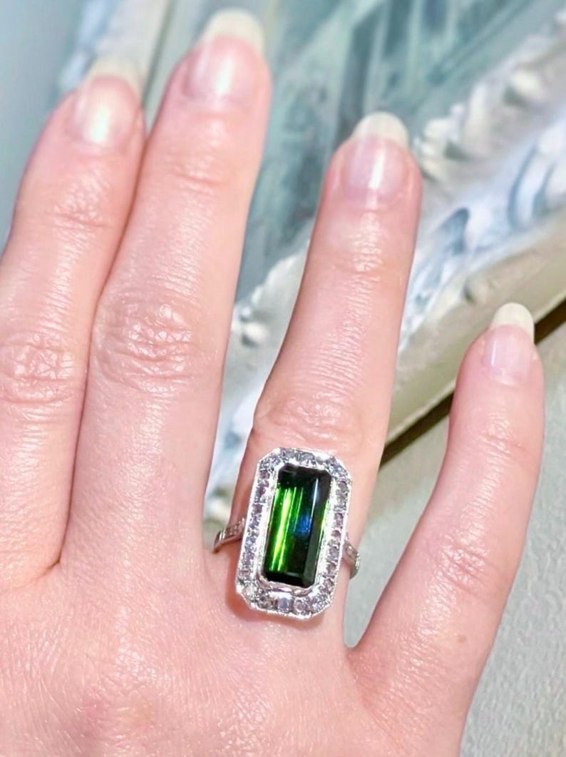 May include: A rectangular emerald-cut green gemstone ring with a halo of small, clear gemstones. The ring is set in a silver-colored band. The ring is on a finger.