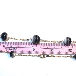 May include: A delicate necklace featuring alternating black and white striped beads strung on a gold-colored chain. The necklace is shown with a pink ruler, with measurements in inches.