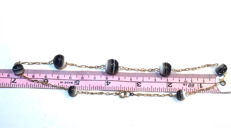 May include: A delicate necklace featuring alternating black and white striped beads strung on a gold-colored chain. The necklace is shown with a pink ruler, with measurements in inches.