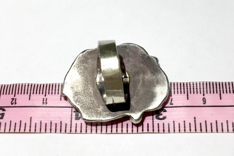 以下が含まれることがあります： テクスチャ加工が施された不規則な形のデザインのシルバーリング。リングはピンクの定規で測定されています。