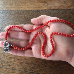 Puede incluir: Un collar de cuentas de coral rojo con un dije de plata en forma de candado. El collar est&aacute; en una mano.
