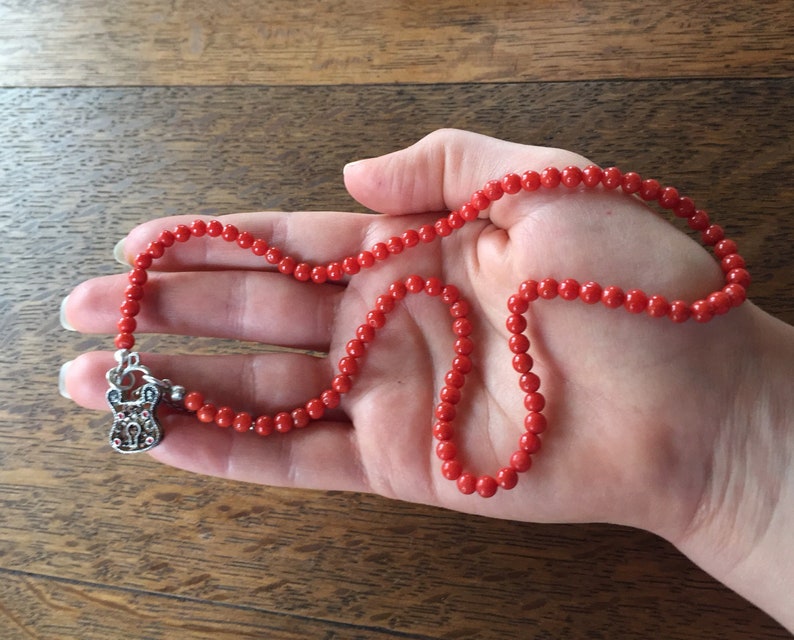 Puede incluir: Un collar de cuentas de coral rojo con un dije de plata en forma de candado. El collar est&aacute; en una mano.
