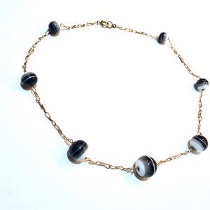 May include: A delicate necklace featuring a gold-toned chain adorned with alternating black and white striped beads. The beads are evenly spaced along the chain, creating a simple yet elegant design. The necklace is displayed on a white background.