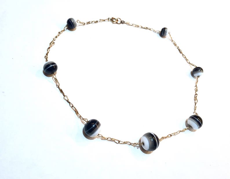 May include: A delicate necklace featuring a gold-toned chain adorned with alternating black and white striped beads. The beads are evenly spaced along the chain, creating a simple yet elegant design. The necklace is displayed on a white background.