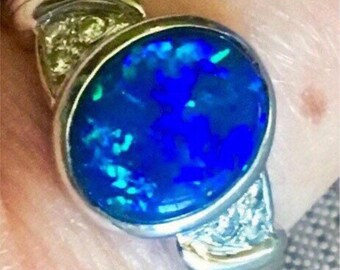 May include: A silver ring featuring a large, oval, deep blue opal gemstone with flashes of green and blue. The ring has a silver band with small accent stones on either side of the opal.