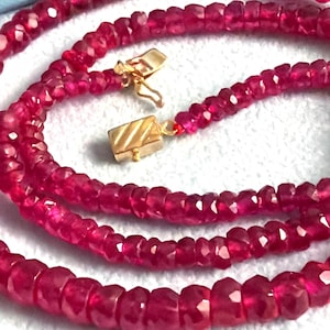 May include: A strand of faceted, deep pinkish-red ruby beads arranged in a circular pattern. The necklace features a rectangular gold-toned clasp with diagonal etched lines and a small hook mechanism, securing the ends of the gemstone strand.