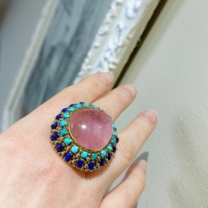May include: A ring with a large pink gemstone surrounded by a halo of blue and turquoise stones.