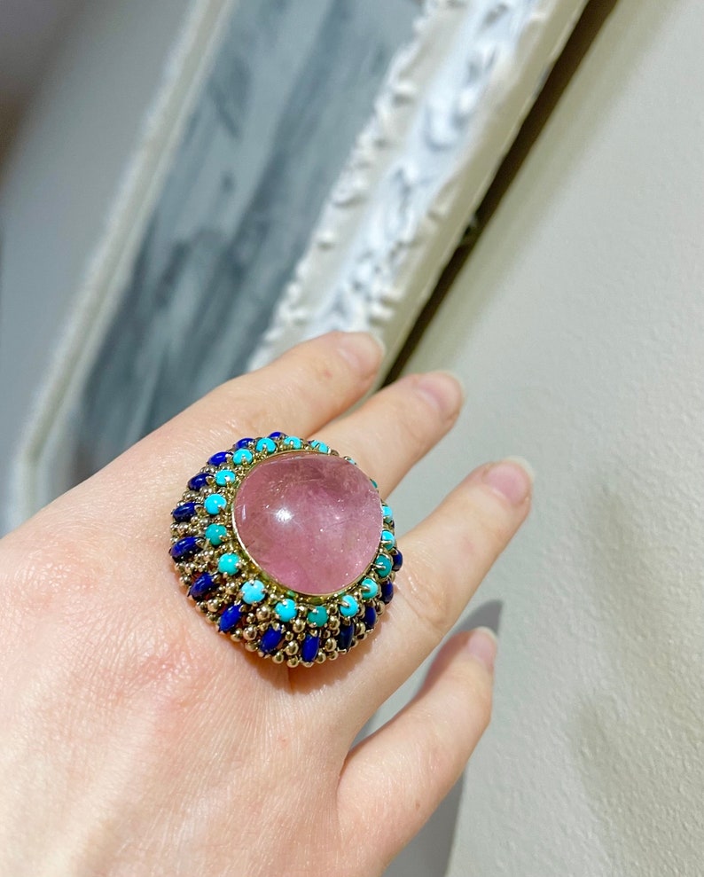 May include: A ring with a large pink gemstone surrounded by a halo of blue and turquoise stones.