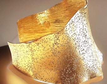 May include: A gold-coloured cuff bracelet with a textured surface. The bracelet has an irregular, flowing shape with a hammered effect and small, dark dots. The edges are uneven, adding to its unique design. The bracelet is a statement piece, suitable for any occasion.