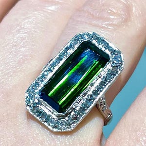 May include: A rectangular ring with a large, vibrant green and blue gemstone. The gemstone is surrounded by a halo of smaller, clear stones set in a silver-colored metal band. The ring is a close-up shot.