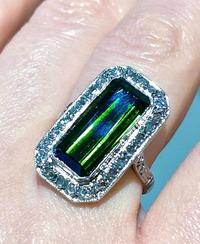 May include: A rectangular ring with a large, vibrant green and blue gemstone. The gemstone is surrounded by a halo of smaller, clear stones set in a silver-colored metal band. The ring is a close-up shot.