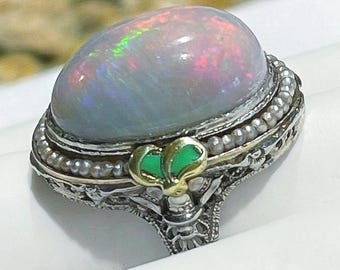 May include: A silver ring featuring a large, oval-shaped cabochon opal with iridescent flashes of red and green. The setting is decorated with a border of tiny white seed pearls, a small gold-toned accent, and intricate metal filigree work.