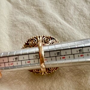 May include: A gold ring with a large red stone set in a filigree design. The ring is being measured with a ring sizer, showing a size 17.