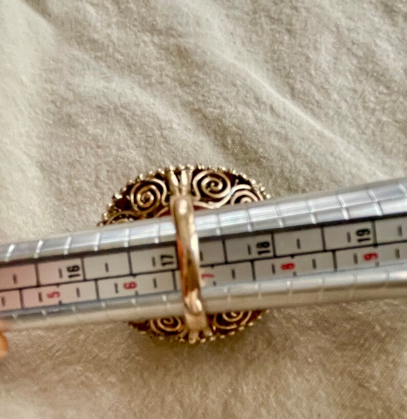 May include: A gold ring with a large red stone set in a filigree design. The ring is being measured with a ring sizer, showing a size 17.