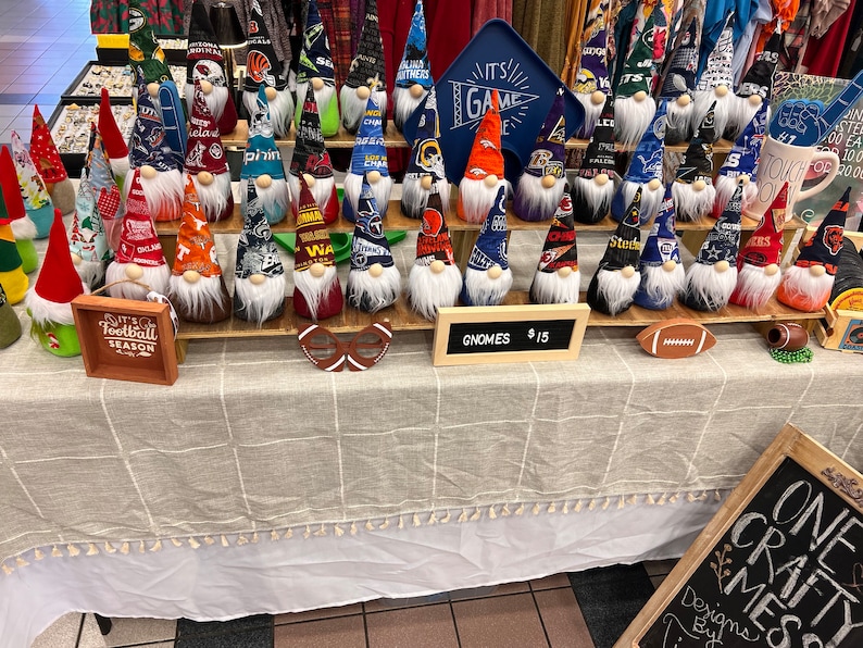May include: A table display of handmade gnomes with various sports team logos and colours. The gnomes are arranged on two shelves and are priced at &pound;15 each. The sign reads "GNOMES &pound;15".