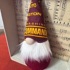 May include: A decorative gnome featuring a tall, pointed burgundy fabric hat with yellow text that reads WASHINGTON COMMANDERS. The gnome has a round, light-coloured wooden nose and a long, thick white faux fur beard covering its burgundy base.