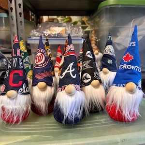 Puede incluir: Siete gnomos que llevan sombreros con diferentes logotipos de equipos de b&eacute;isbol. Los gnomos llevan todos barbas blancas y tienen cuerpos rojos, azules o verdes. Los logotipos incluyen los Cleveland Guardians, los Washington Nationals, los Atlanta Braves, los New England Patriots, los Seattle Mariners, los Toronto Blue Jays y los Los Angeles Dodgers.