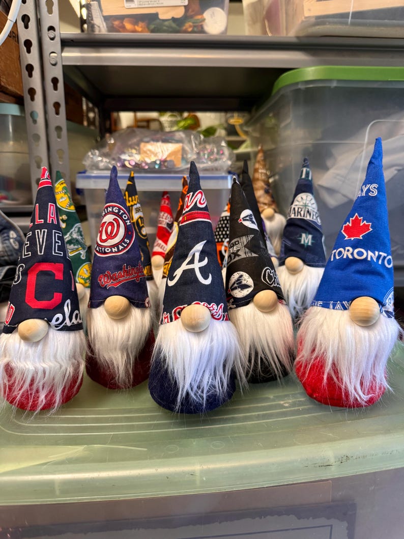 Puede incluir: Siete gnomos que llevan sombreros con diferentes logotipos de equipos de b&eacute;isbol. Los gnomos llevan todos barbas blancas y tienen cuerpos rojos, azules o verdes. Los logotipos incluyen los Cleveland Guardians, los Washington Nationals, los Atlanta Braves, los New England Patriots, los Seattle Mariners, los Toronto Blue Jays y los Los Angeles Dodgers.