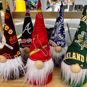 Puede incluir: Cinco figuras de gnomos con diferentes logotipos de equipos de b&eacute;isbol. Los gnomos llevan sombreros con los logotipos de los Atlanta Braves, los St. Louis Cardinals, los New York Mets, los Oakland Athletics y un equipo de b&eacute;isbol gen&eacute;rico. Los gnomos llevan todos barbas blancas y tienen narices de madera.