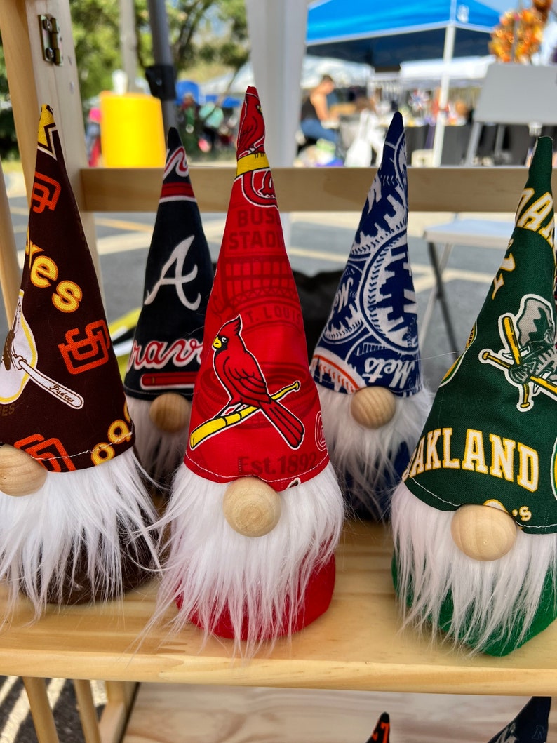 Puede incluir: Cinco figuras de gnomos con diferentes logotipos de equipos de b&eacute;isbol. Los gnomos llevan sombreros con los logotipos de los Atlanta Braves, los St. Louis Cardinals, los New York Mets, los Oakland Athletics y un equipo de b&eacute;isbol gen&eacute;rico. Los gnomos llevan todos barbas blancas y tienen narices de madera.