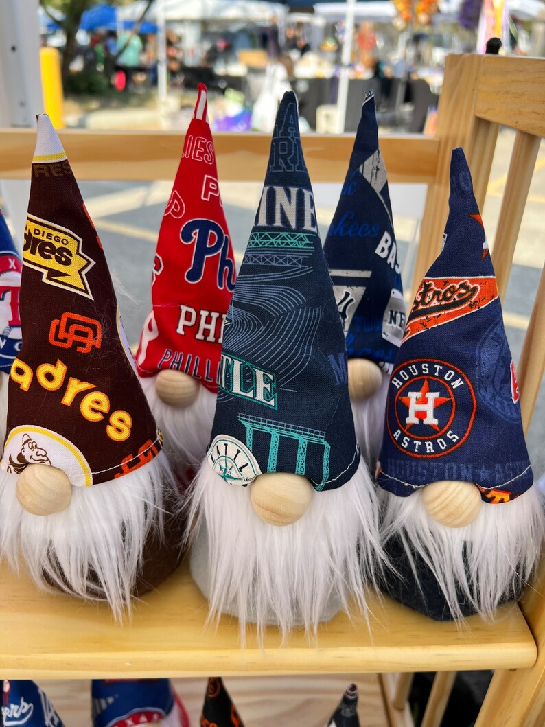 Puede incluir: Cinco gnomos con barbas blancas y narices de madera. Cada gnomo lleva un sombrero diferente con el logotipo de un equipo deportivo. Los sombreros son marrones con el logotipo de los San Diego Padres, rojos con el logotipo de los Philadelphia Phillies, azules con el logotipo de los New York Mets, azules con el logotipo de los Kansas City Royals y azules con el logotipo de los Houston Astros.