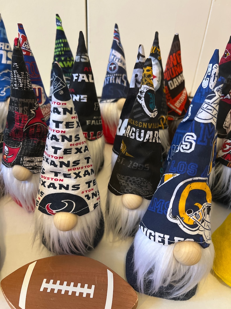 May include: A collection of 12 gnomes featuring different NFL team logos. The gnomes are made of fabric and have white beards. The logos include the Atlanta Falcons, Houston Texans, Jacksonville Jaguars, Los Angeles Rams, and Tennessee Titans.
