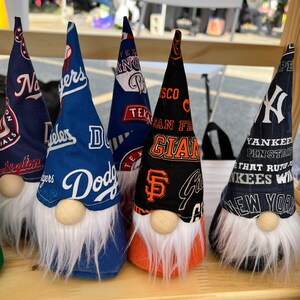 Puede incluir: Cinco gnomos con diferentes logotipos de equipos de b&eacute;isbol en sus sombreros. Los gnomos est&aacute;n alineados en un estante de madera. Los logotipos son para los New York Yankees, los San Francisco Giants, los Los Angeles Dodgers, los Texas Rangers y los Washington Nationals.