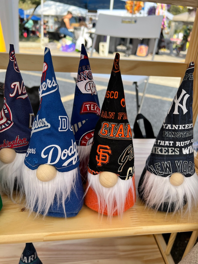 Puede incluir: Cinco gnomos con diferentes logotipos de equipos de b&eacute;isbol en sus sombreros. Los gnomos est&aacute;n alineados en un estante de madera. Los logotipos son para los New York Yankees, los San Francisco Giants, los Los Angeles Dodgers, los Texas Rangers y los Washington Nationals.